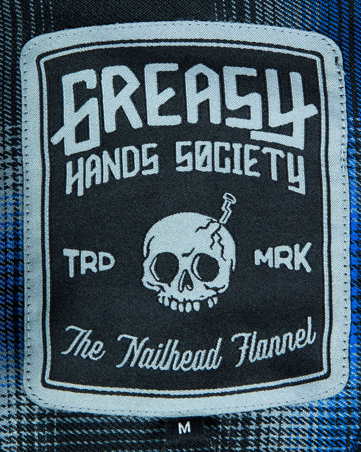 tag Patch on the nailhead blue and black flannel with 'Greasy Hands Society the nailhead flannel' text design with a skull on a flannel fabric background - The Nailhead Flannel by Greasy Hands Society