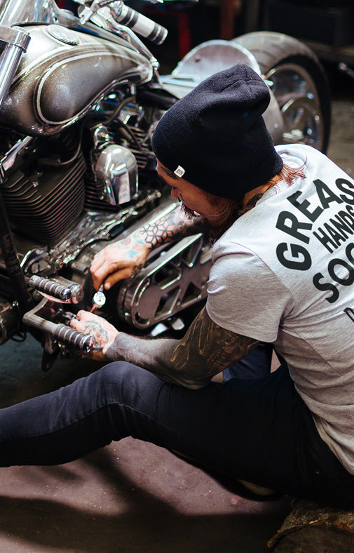 Person working on a motorcycle wearing a t-shirt with text while wearing a greasy hands society beanie