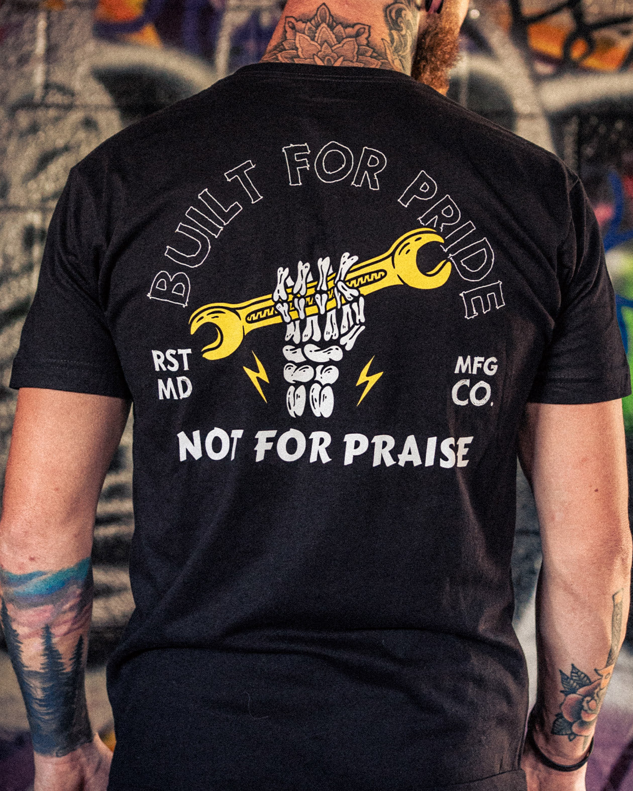 Person wearing a black t-shirt with a graphic design and text on the back, standing in front of a graffiti-covered wall - Built for Pride men's black graphic t-shirt by greasy hands society
