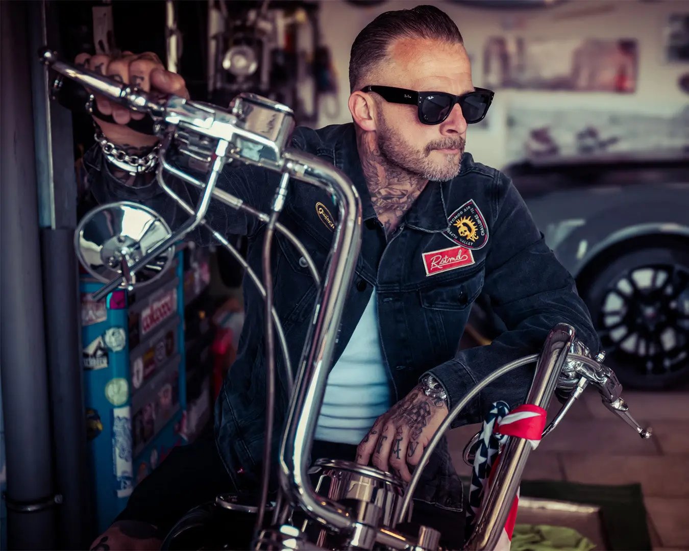 man riding a motorcycle with tattoos and sunglasses wearing a denim jacket with patched that say 'RSTMD' - The Black Denim Jacket by Greasy Hands Society