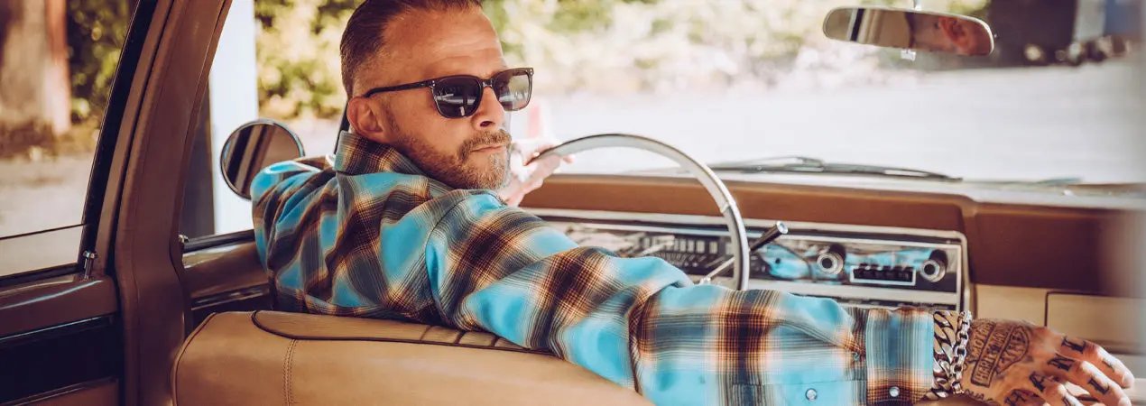 A man wearing a blue and brown plaid flannel shirt and black sunglasses sits in the driver's seat of a vintage car, holding the steering wheel - Cold Start Flannel by Greasy Hands Society