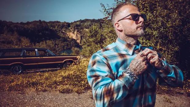 A man wearing a blue and brown plaid button-down shirt and sunglasses stands outdoors in front of a vintage car and hills, adjusting his collar - Cold Start Flannel by Greasy Hands Society