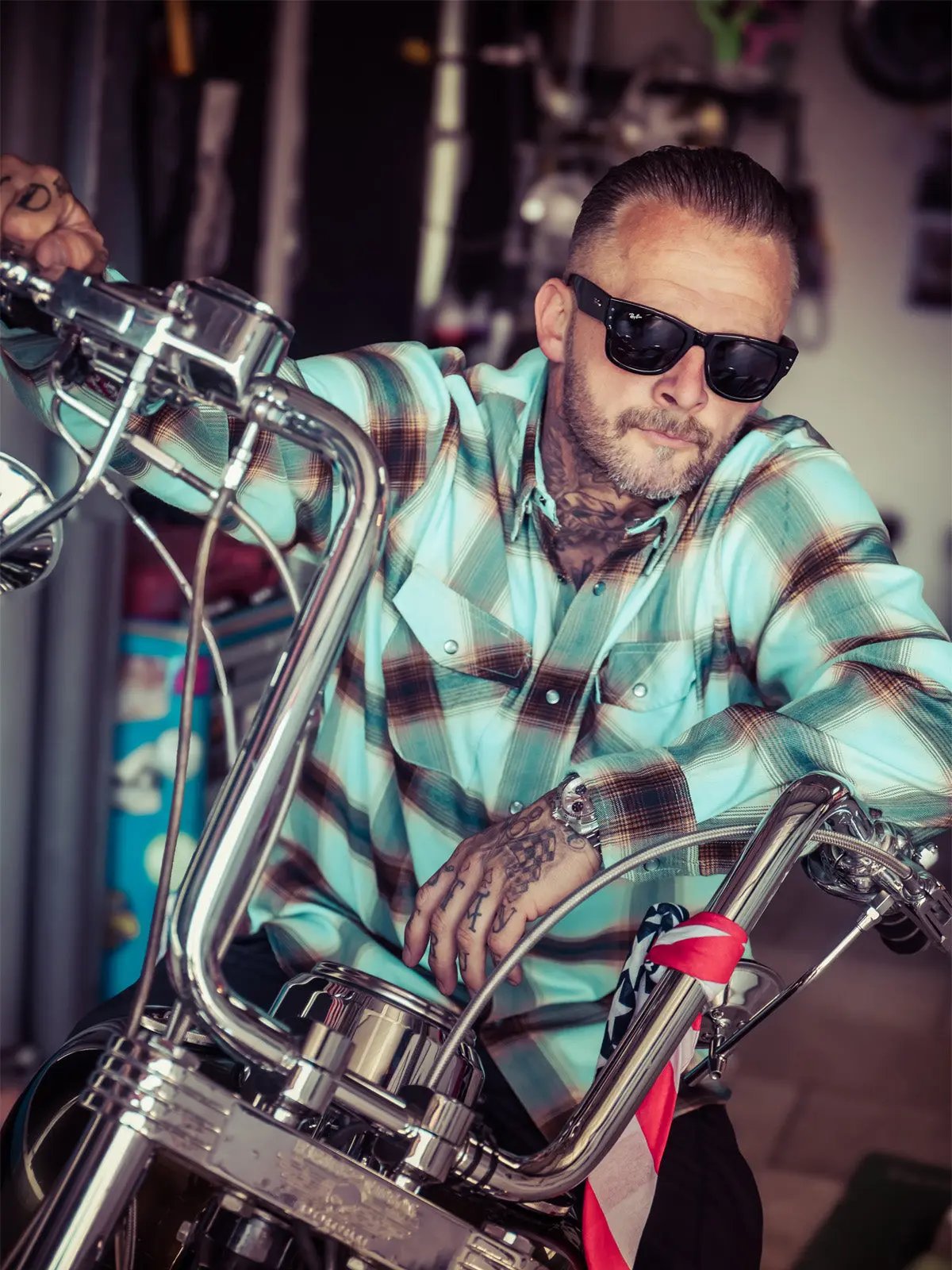 A man with tattoos and sunglasses wearing a light blue and brown plaid button-down shirt, leaning on the chrome handlebars of a motorcycle in a garage setting - Cold Start Flannel by Greasy Hands Society