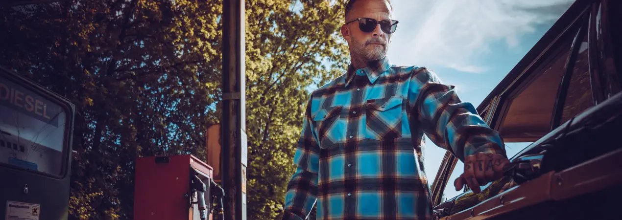 A man wearing a blue and brown plaid button-down flannel shirt and sunglasses stands next to an open car door in an outdoor setting with trees and a cloudy sky - Cold Start Flannel by Greasy Hands Society