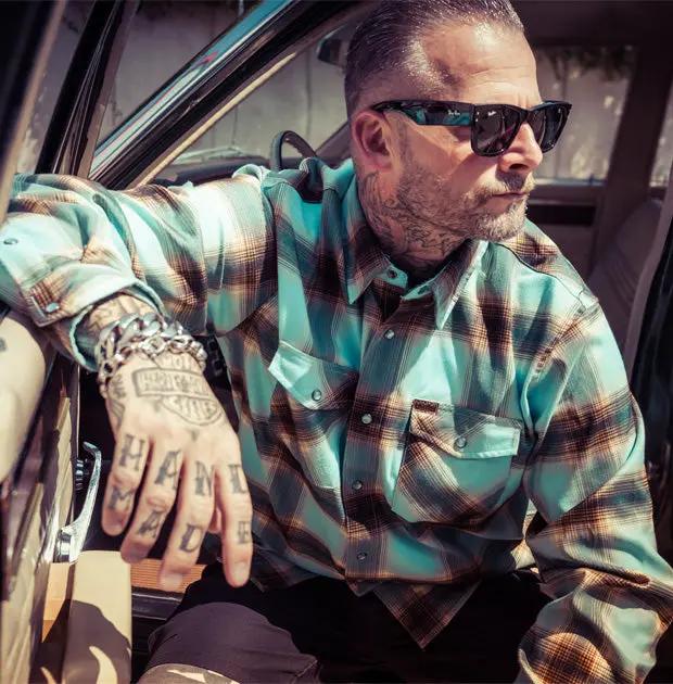 A man with tattoos and sunglasses leans in a car, wearing a blue and brown plaid button-down flannel shirt - Cold Start Flannel by Greasy Hands Society
