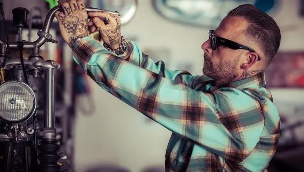 A tattooed man wearing black sunglasses and a blue, brown, and beige plaid flannel shirt works on a motorcycle in a garage - Cold Start Flannel by Greasy Hands Society
