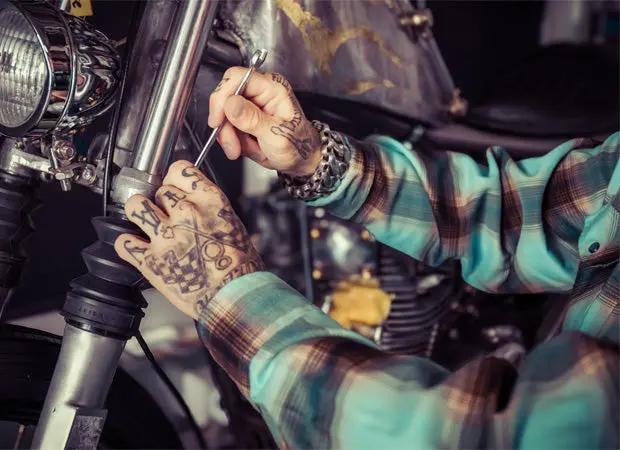 A close-up lifestyle shot of a tattooed person in a teal and brown plaid shirt working on a motorcycle, using a wrench on the front - Cold Start Flannel by Greasy Hands Society