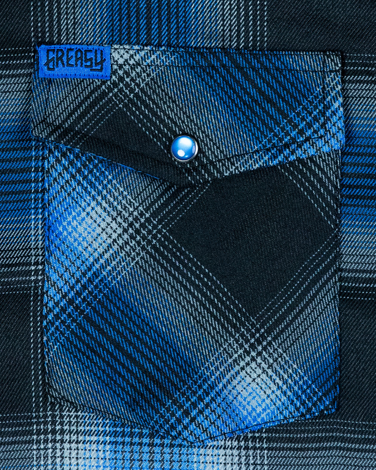 Close-up of a blue and black plaid flannel shirt pocket with a button, featuring the brand 'Greasy' - called the nailhead flannel.