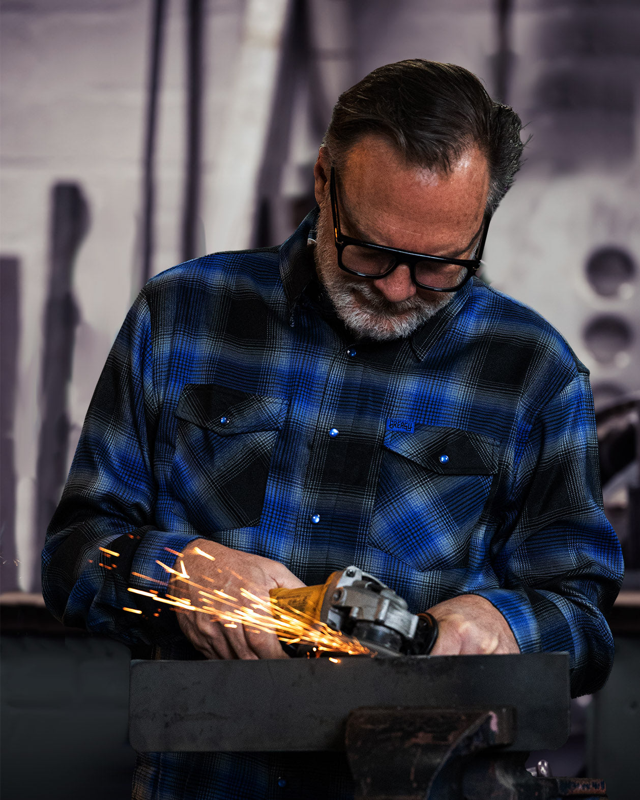 Man wearing a blue plaid shirt using a grinder with sparks in a workshop setting.