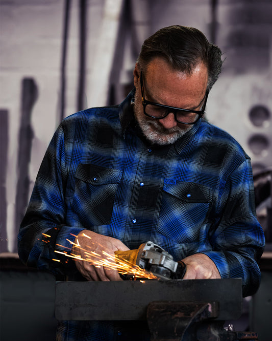 Man wearing a blue plaid shirt using a grinder with sparks in a workshop setting.