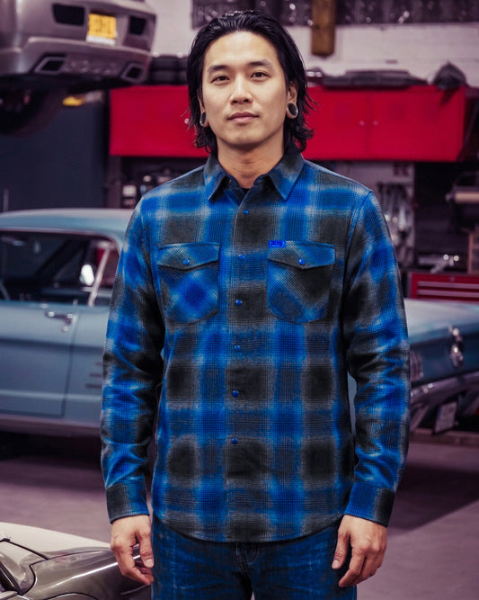 Man wearing a blue and gray plaid flannel shirt standing in a garage with cars in the background - The Nailhead Flannel by Greasy Hands Society