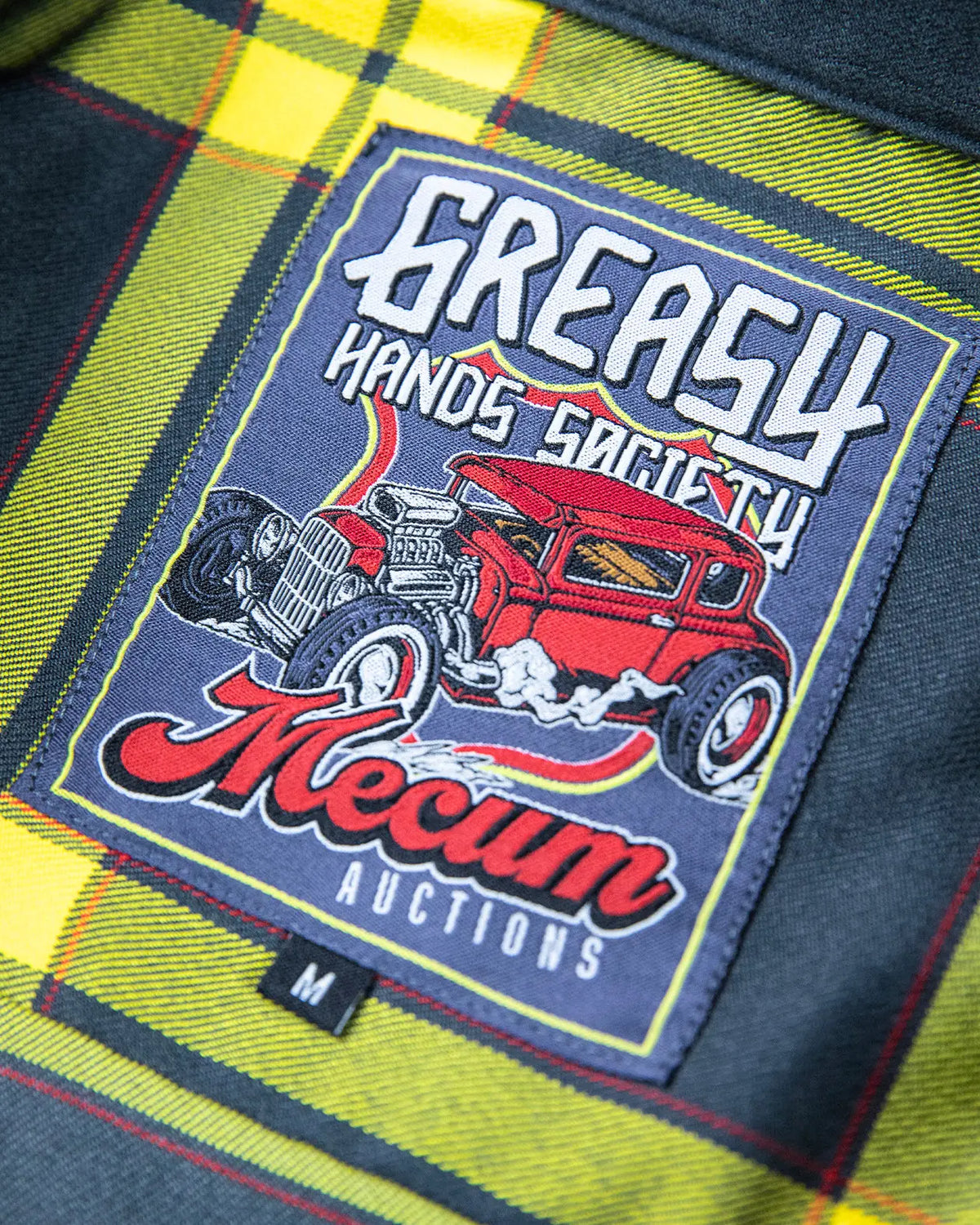 A close-up of a rectangular embroidered patch on a plaid shirt. The patch features a red hot rod car and the text 'GREASY HANDS SOCIETY' and 'Mecum AUCTIONS' - The Main Attraction Flannel - Mecum Auctions x Greasy Hands Society Collab Flannel