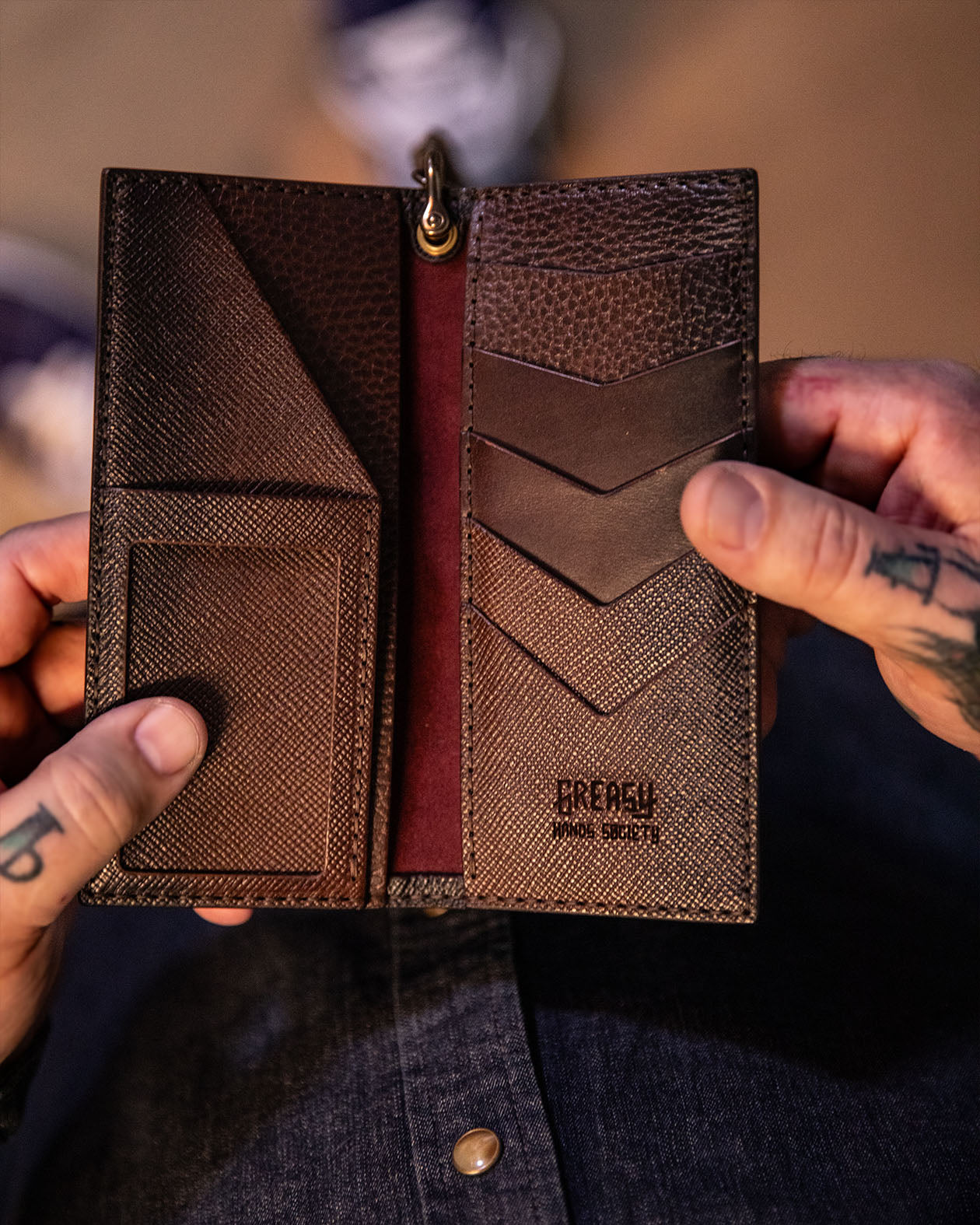 black leather wallet held open by a person with visible tattoos, brand 'Greasy' on the wallet.