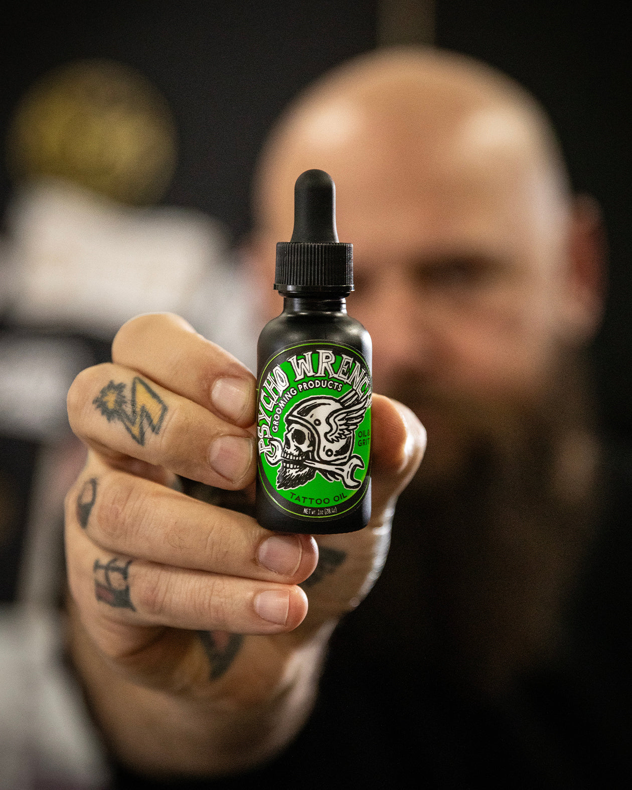 man holding up a bottle of psycho wrench oil & grit grooming products tattoo oil with a blurred background