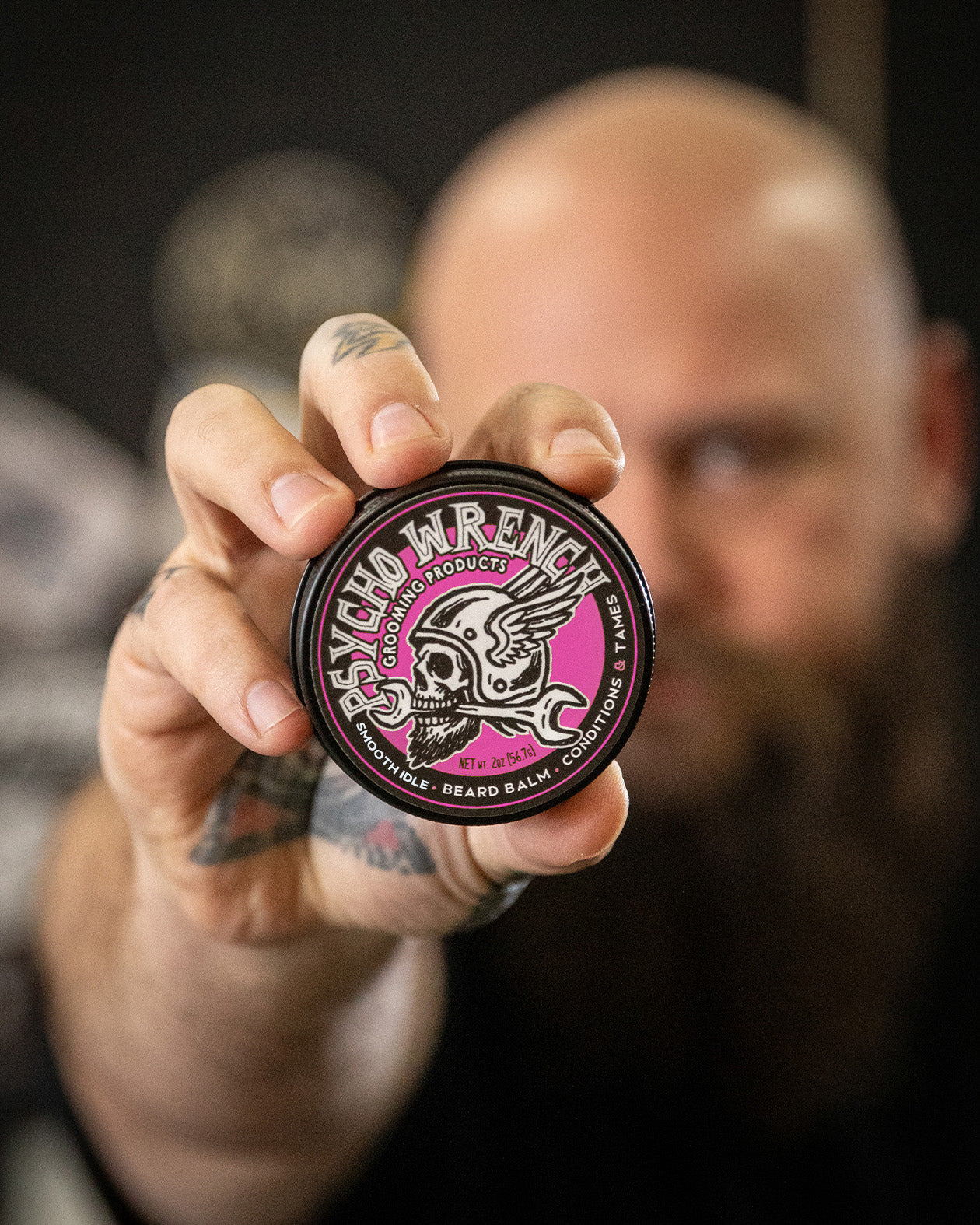Man holding a pink and black container of psycho wrench grooming product beard balm 2oz with a logo, blurred background