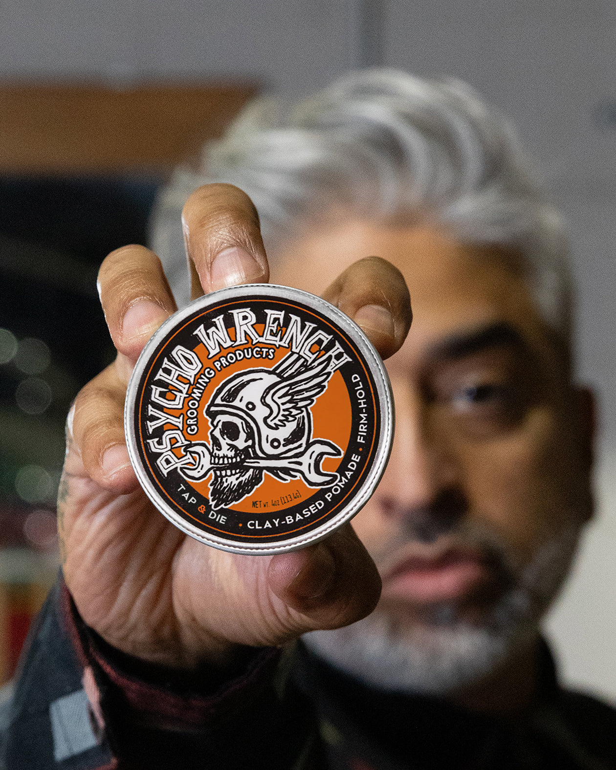 Person holding a product container with a brand logo against a blurred background - Psycho Wrench Tap & Die Clay Pomade by Greasy Hands Society