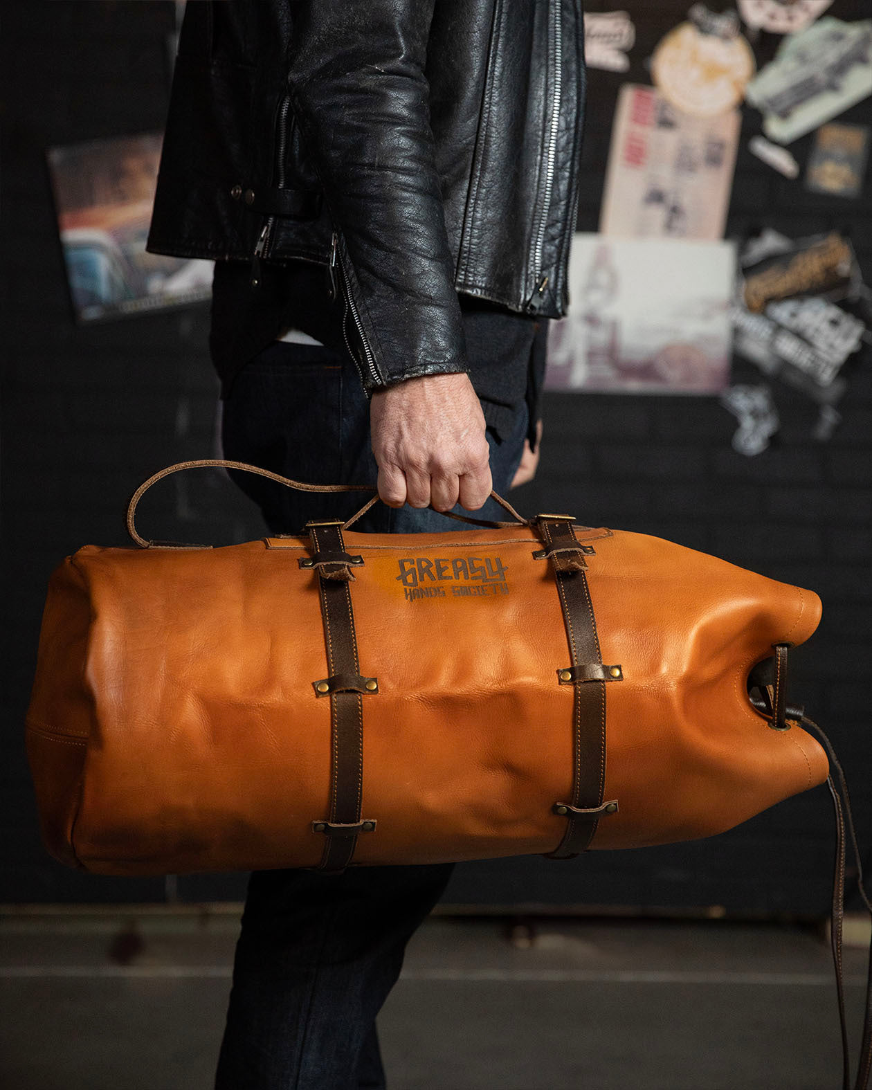 Person holding a brown leather duffel bag with 'Greasy Hands Society' branding against a dark background.