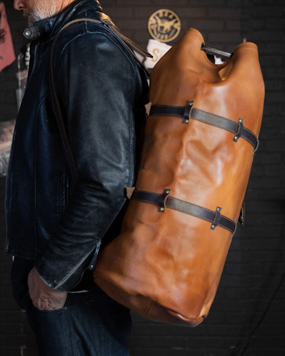 Person wearing a large brown leather bag with dark brown straps on a dark background from greasy hands society