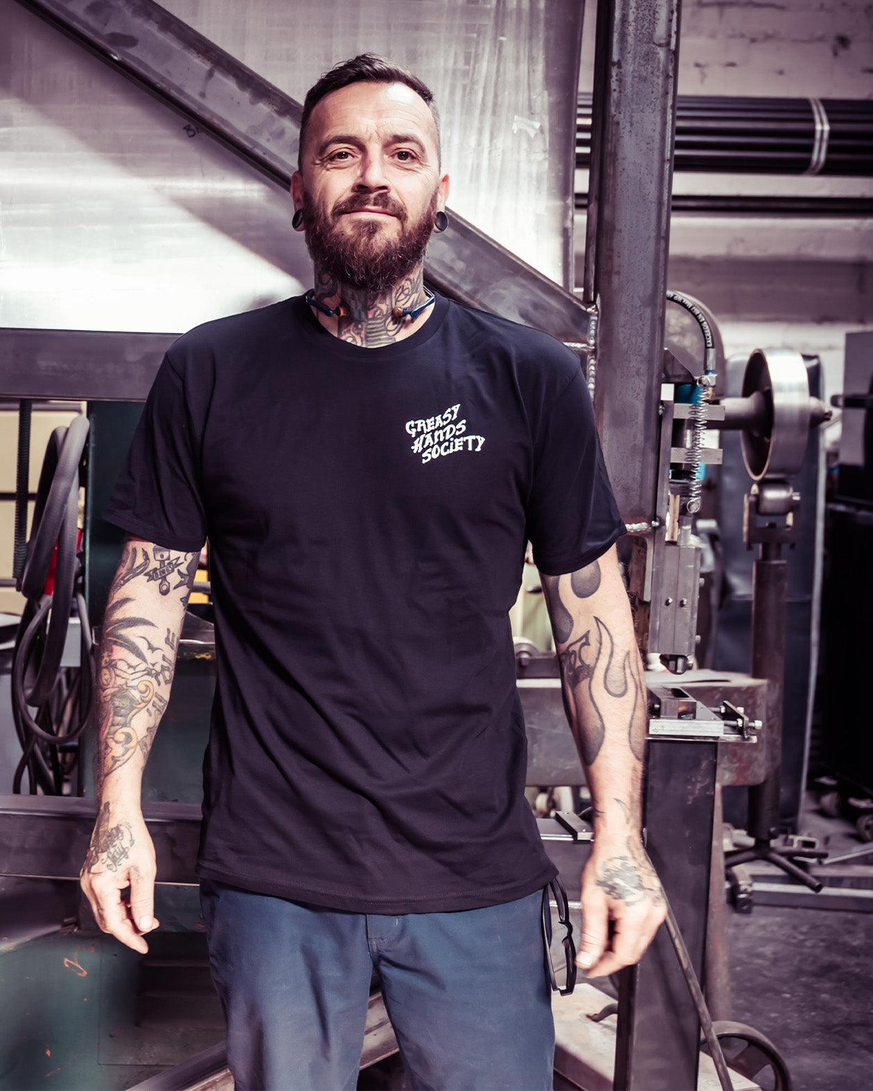 Man with tattoos wearing a black t-shirt that says 'greasy hands society' on the left chest in an industrial setting