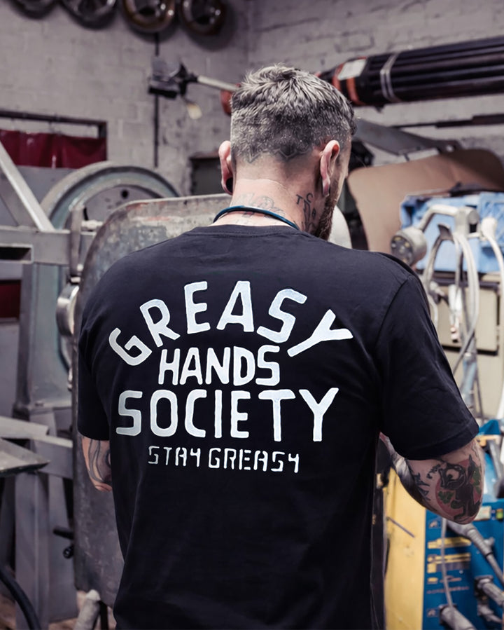 Person wearing a black t-shirt with 'Greasy Hands Society Stay Greasy' text in a workshop setting - Factory Sealed mens graphic tee by Greasy Hands Society