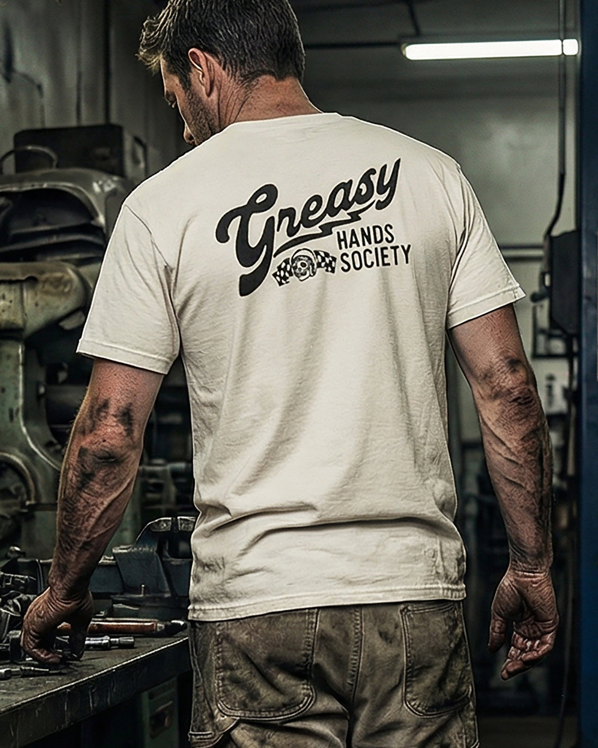 Person wearing a beige 'Greasy Hands Society' t-shirt called the destination tee in a workshop setting