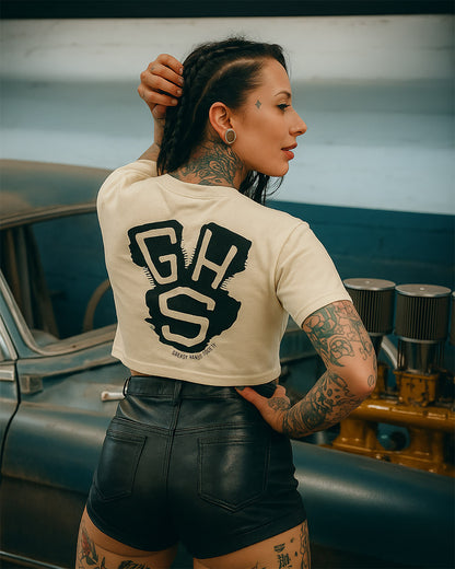 woman wearing a white t-shirt with 'GHS' on the back with an engine sillhouetted in the back and leather shorts, standing next to a vintage car.
