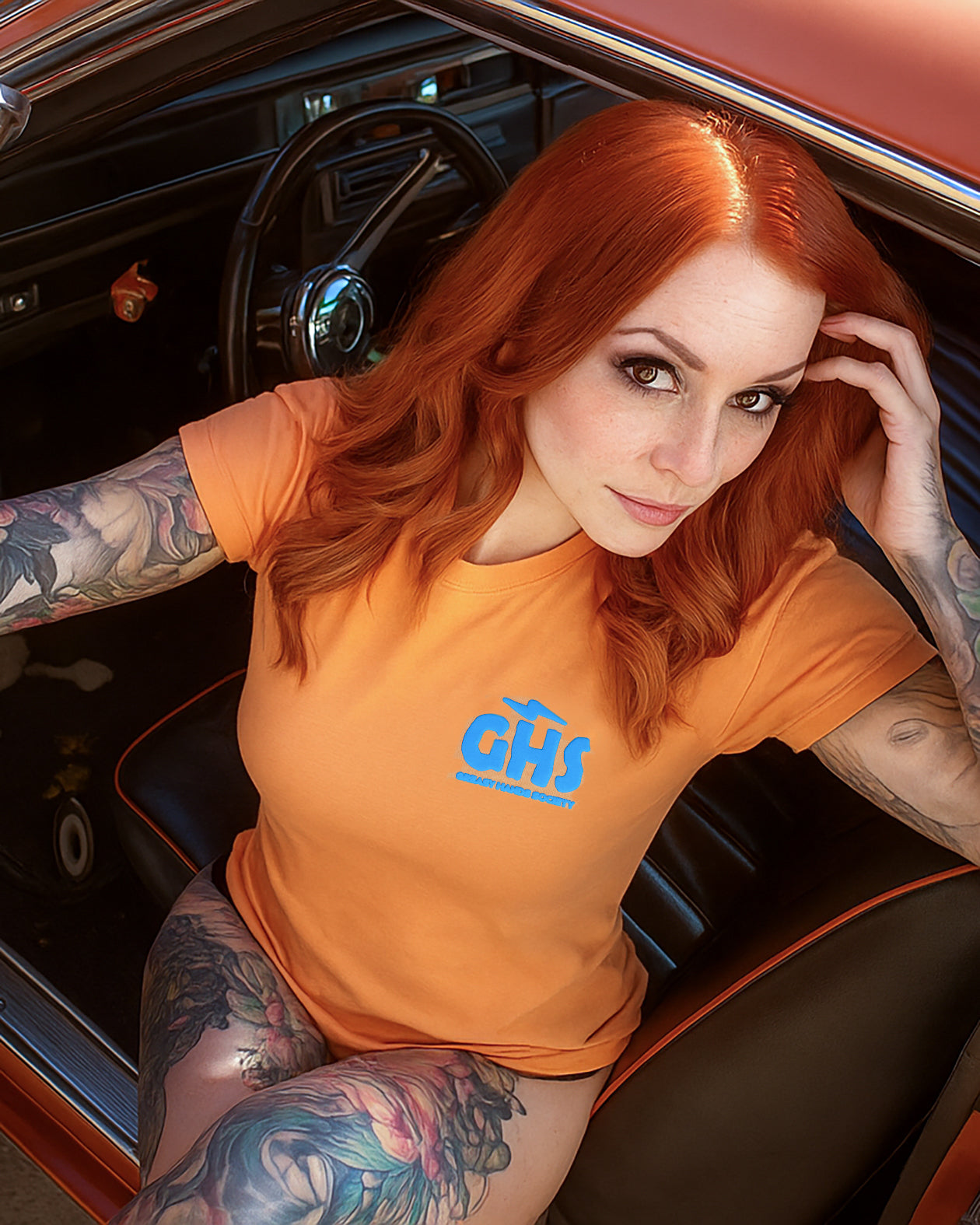 women's go faster graphic tee - Woman with tattoos wearing an orange t-shirt with a blue GHS on the left chest inside a classic car.
