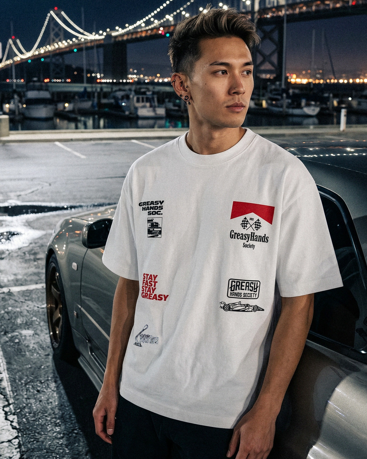 Person wearing a white boxy streetwear JDM t-shirt with logos in front of a car and bridge at night.