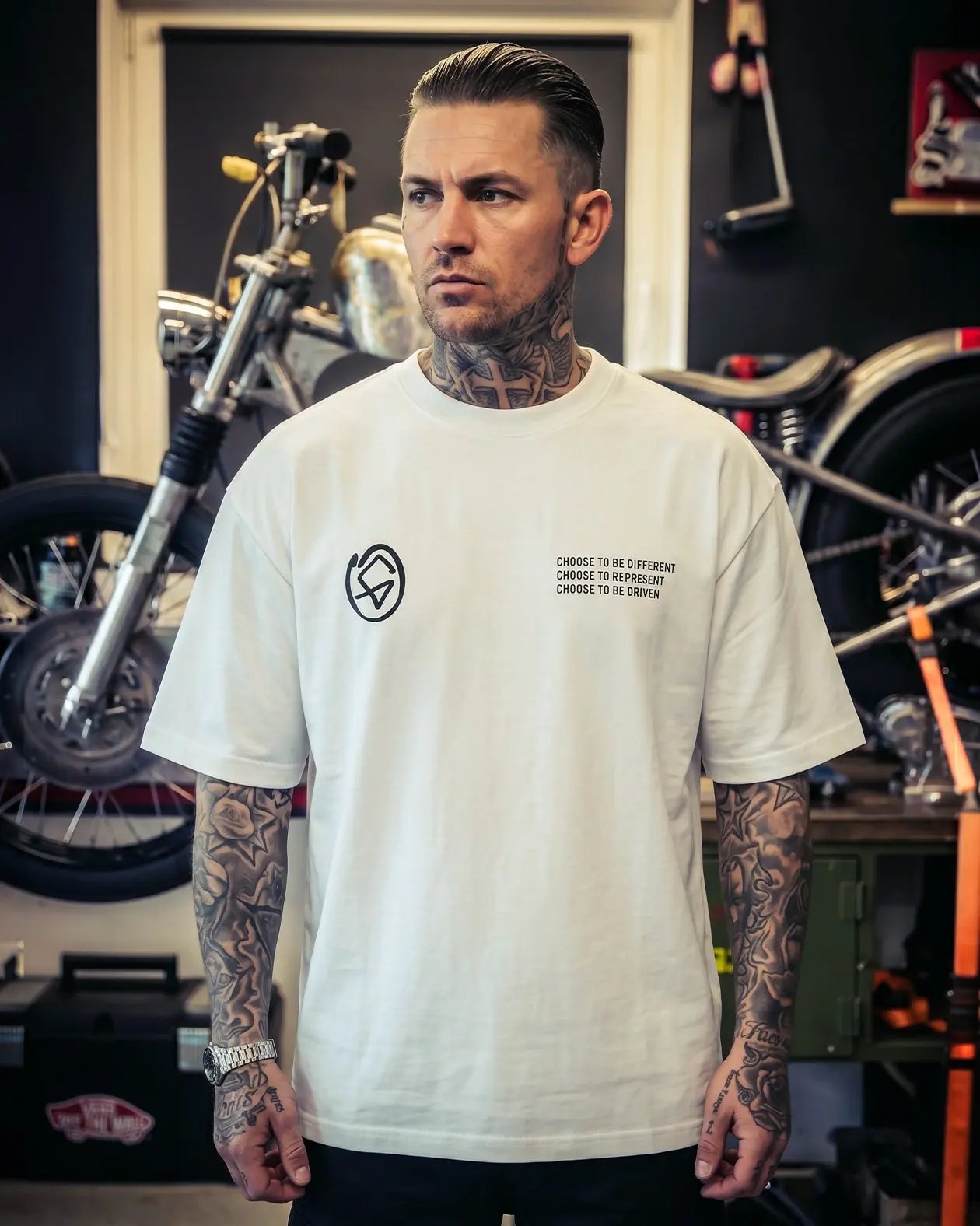 Man wearing a white t-shirt with a logo and text in a garage setting with motorcycles.- Slam'd White Heavyweight graphic Tee by Greasy Hands Society