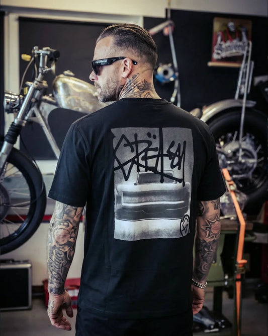 Person wearing a black t-shirt with graphic design in a motorcycle-themed room - The Tailgate Black automotive graphic tee by Greasy Hands Society