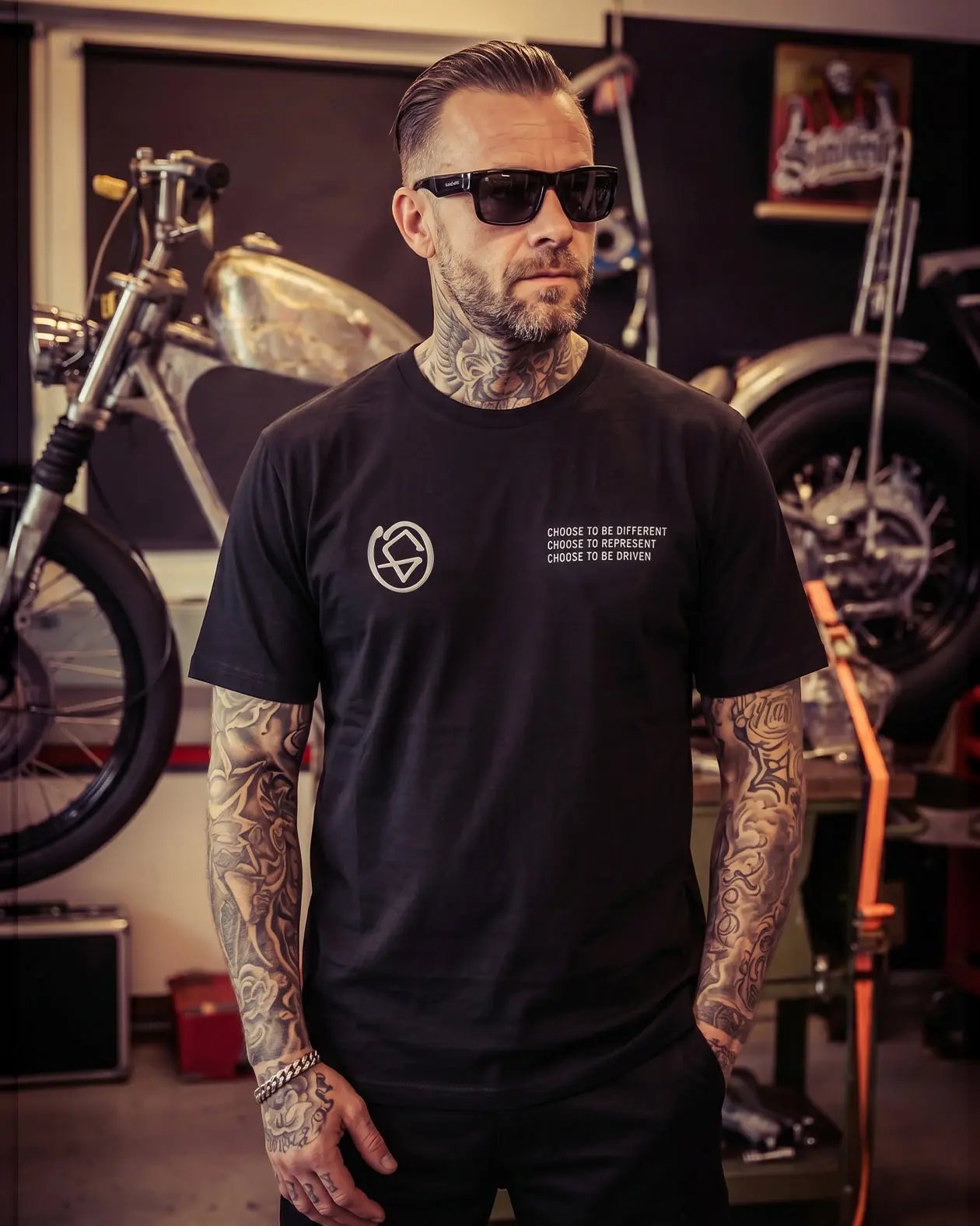 Man wearing a black t-shirt with a logo and text in a garage setting - The Tailgate Black automotive graphic tee by Greasy Hands Society