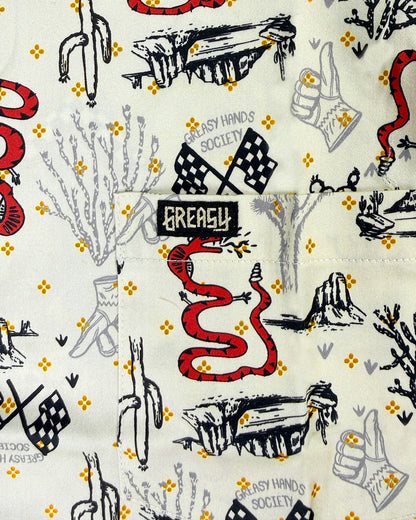 Close-up of a Desert patterned printed fabric featuring a red snake, black and gray cactus and rock illustrations, checkered flags, thumbs-up hands, and small yellow diamond motifs on an off-white background. A black fabric label reading 'GREASY' is sewn into the design - Baja Button-Up Shirt by Greasy Hands Society