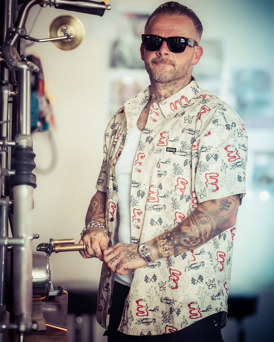A man wearing black sunglasses and a short-sleeve button-up shirt with a red, black, and white graphic print, standing in a workshop environment -The Baja Button-Up Shirt by Greasy Hands Society