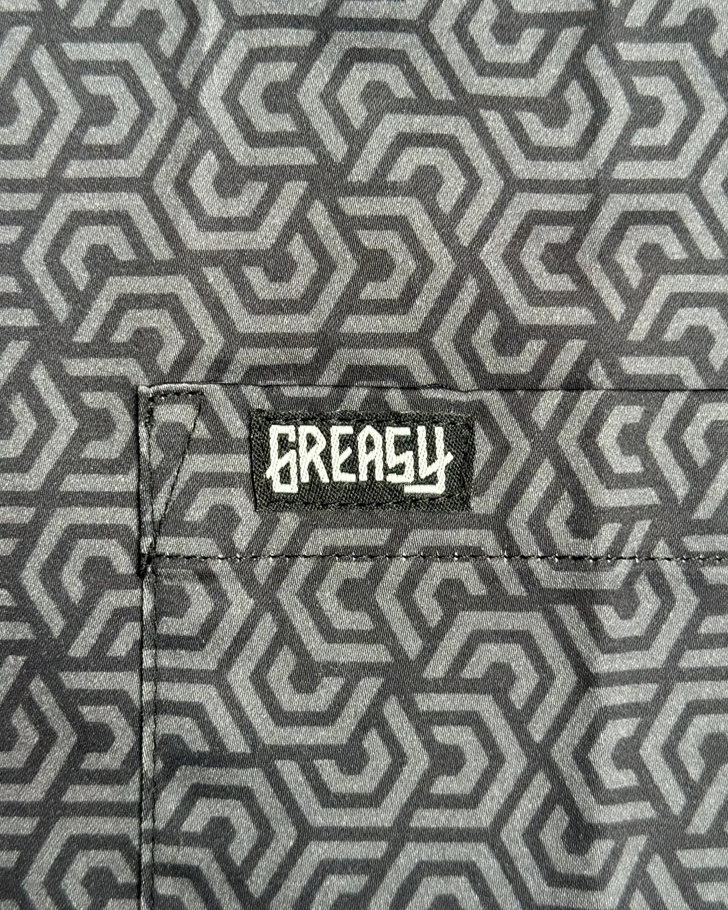 Close-up of a gray and black geometric patterned fabric with a black patch featuring the white 'GREASY' brand logo on the pocket - The Burnout Button-Up Shirt by Greasy Hands Society