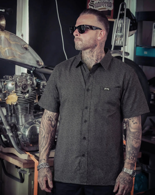 A tattooed man wearing a dark gray short-sleeve button-up shirt with a subtle geometric pattern, black sunglasses, and a watch, standing in a garage setting next to a motorcycle engine - The Burnout Button-Up Shirt by Greasy Hands Society