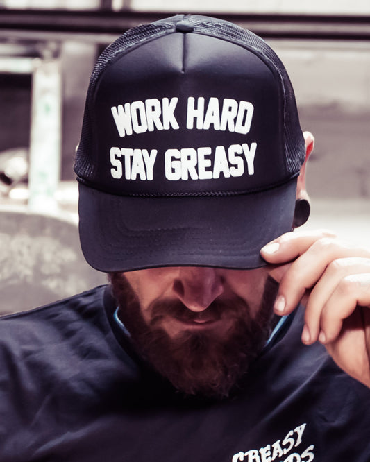 work hard stay greasy black  foam trucker hat with white text on a man who is holding the brim of the hat