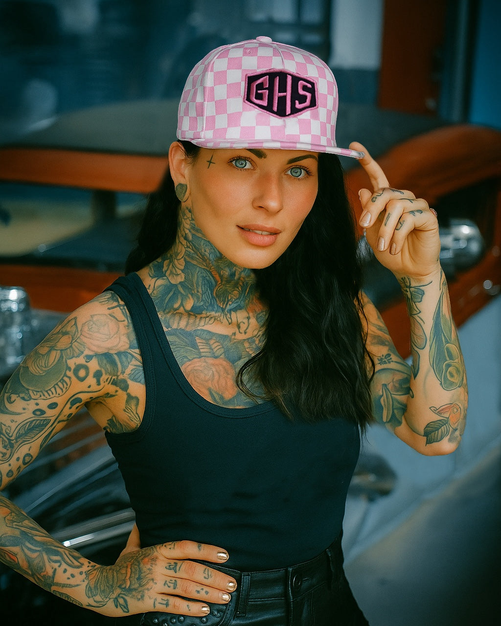 Woman wearing a pink checkerboard hat with a GHS badge, tattoos on arms and neck, black top, and leather shorts.