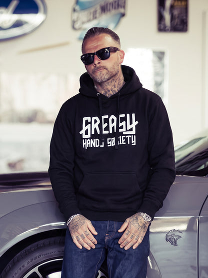A man wearing a black hoodie with 'GREASY HANDS SOCIETY' printed in white on the front, standing next to a gray car in a garage setting - GHS Logo Hoodie by Greasy Hands Society
