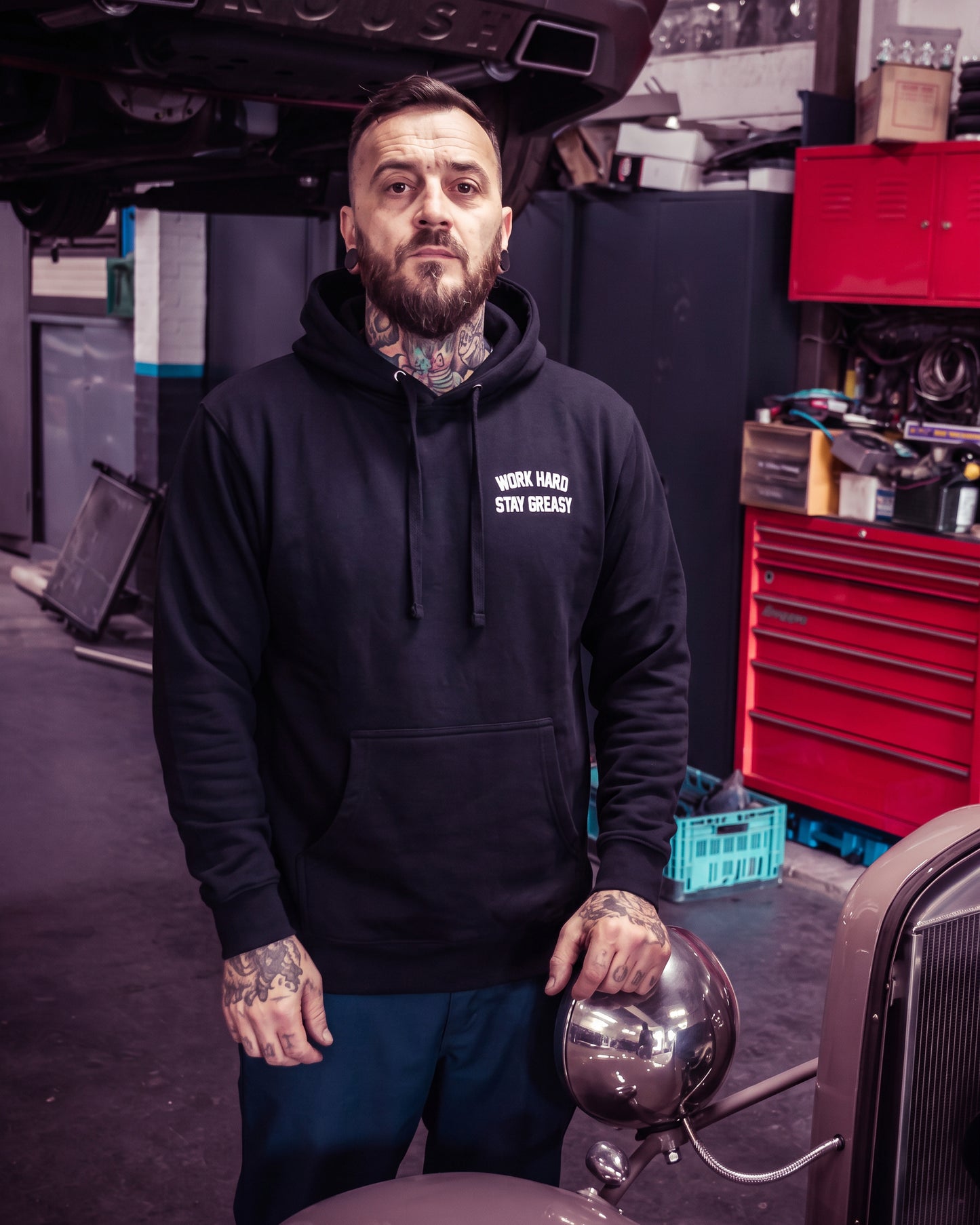 A man with a beard and tattoos wearing a black hoodie with the white text 'WORK HARD STAY GREASY' stands in an auto repair shop - Work Hard Stay Greasy Hoodie by Greasy Hands Society