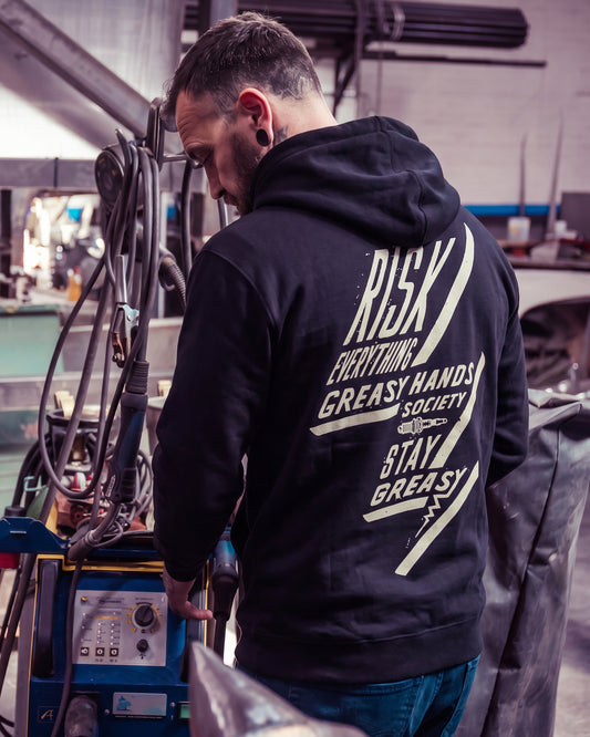 A man wearing a black hoodie with a large white graphic print on the back, standing in a workshop environment - The Bolted Down Hoodie by Greasy Hands Society