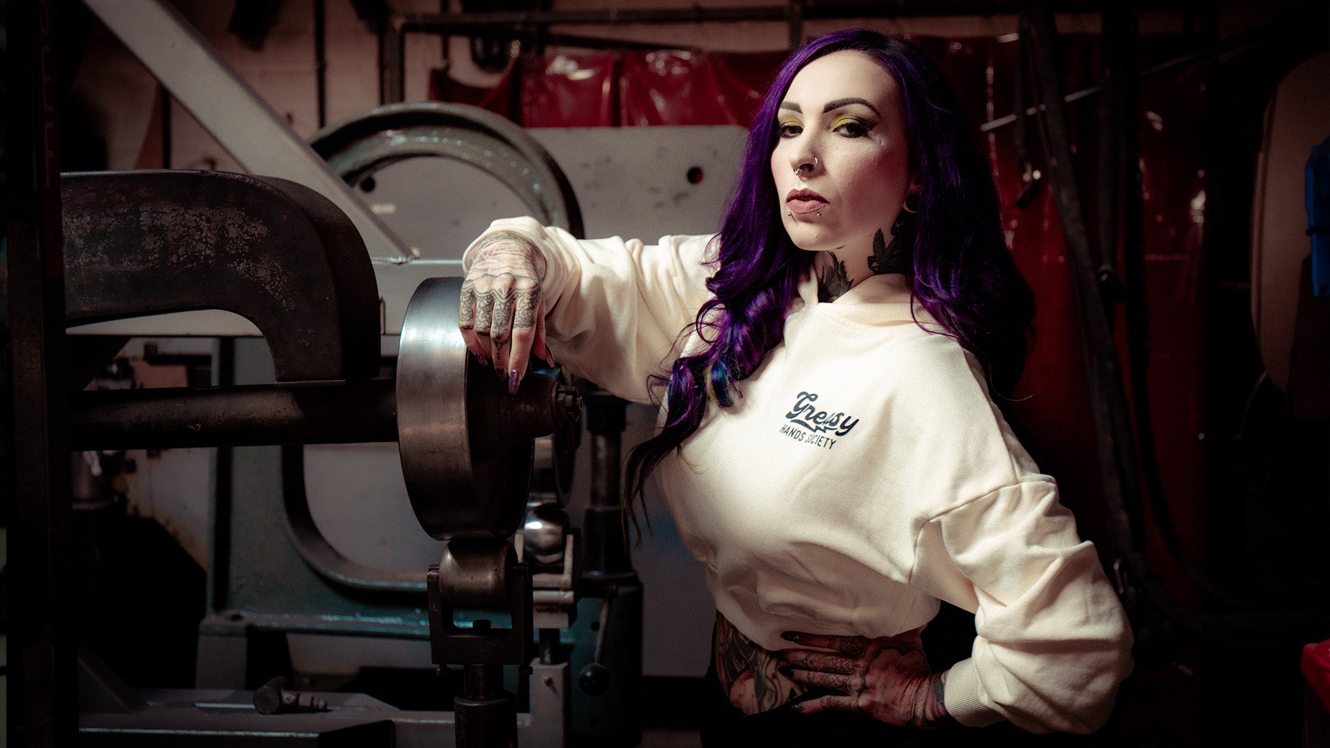 Person with purple hair wearing a white sweatshirt that says 'greasy hands society" in a workshop setting