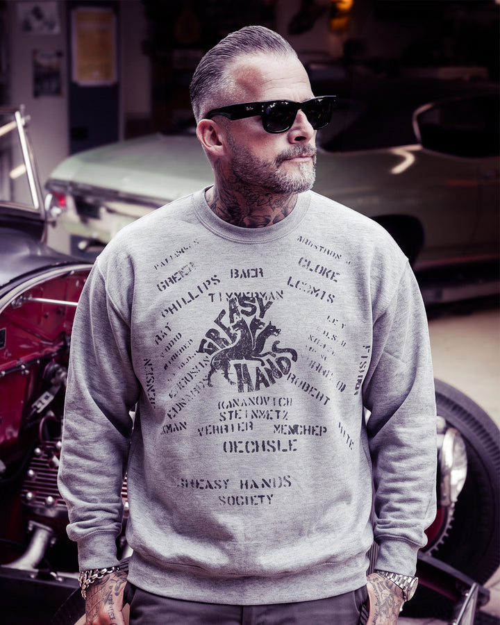 A man wearing a light gray crewneck sweatshirt with a black graphic print that lists alot of names of navy warriors ' and 'Greasy Hands Society', standing in front of a vintage car and motorcycle in a garage - Navy Dogs Crewneck Sweatshirt by Greasy Hands Society