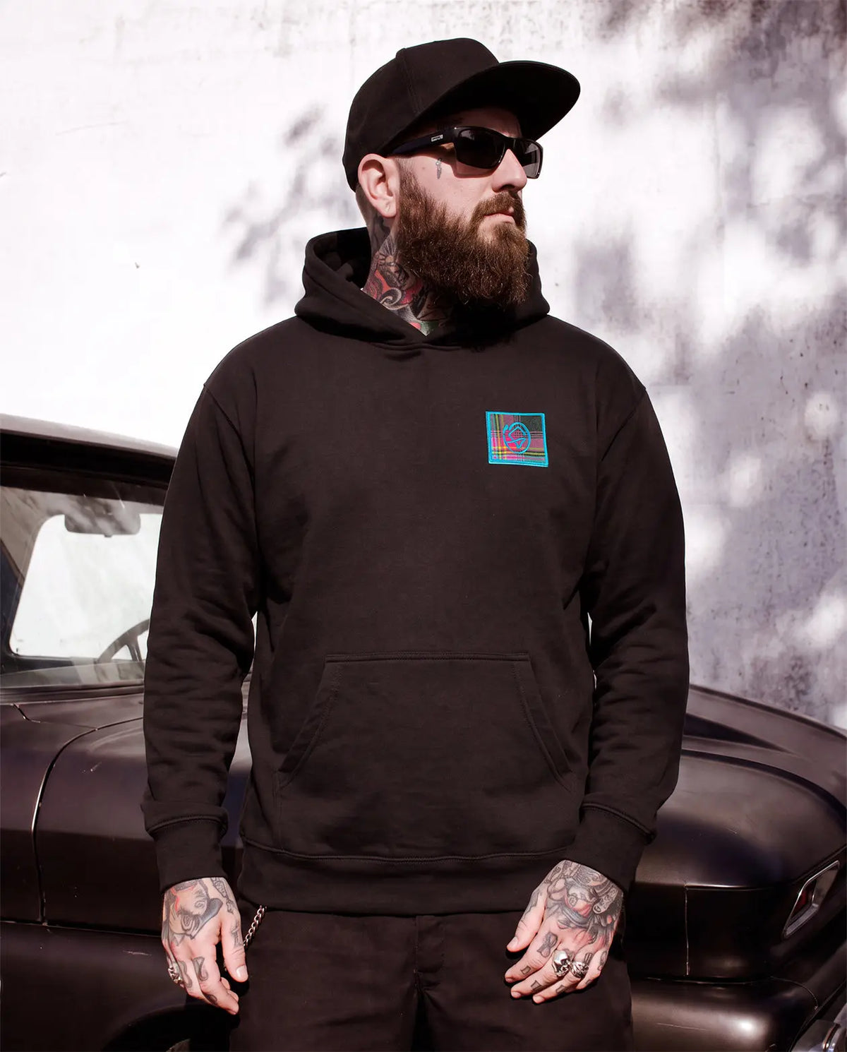 A man with a beard, sunglasses, and tattoos wearing a black hoodie with a colorful square graphic on the left chest, standing in front of a vintage car and textured wall - Blitzkreig Hoodie by Greasy Hands Society 