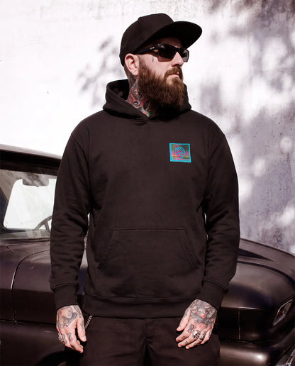 A man with a beard, sunglasses, and tattoos wearing a black hoodie with a colorful square graphic on the left chest, standing in front of a vintage car and textured wall - Blitzkreig Hoodie by Greasy Hands Society 