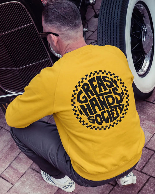 A man wearing a mustard yellow sweatshirt with a large black circular graphic reading 'Greasy Hands Society' on the back, crouching beside a classic car on a paved surface - The Slicks Sweatshirt by Greasy Hands Society