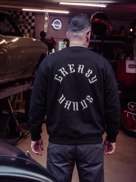 A man viewed from behind wearing a black crewneck sweatshirt with 'GREASY HANDS' printed in white gothic letters on the back, standing in a garage setting - Roller Crew Sweatshirt by Greasy Hands Society