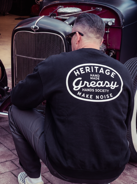 A man wearing a black sweatshirt with a large white circular graphic on the back, crouching in front of a vintage car. The sweatshirt features the text 'HERITAGE - HAND MADE - Greasy - HANDS SOCIETY -MAKE NOISE' - The Heritage Sweatshirt by Greasy Hands Society