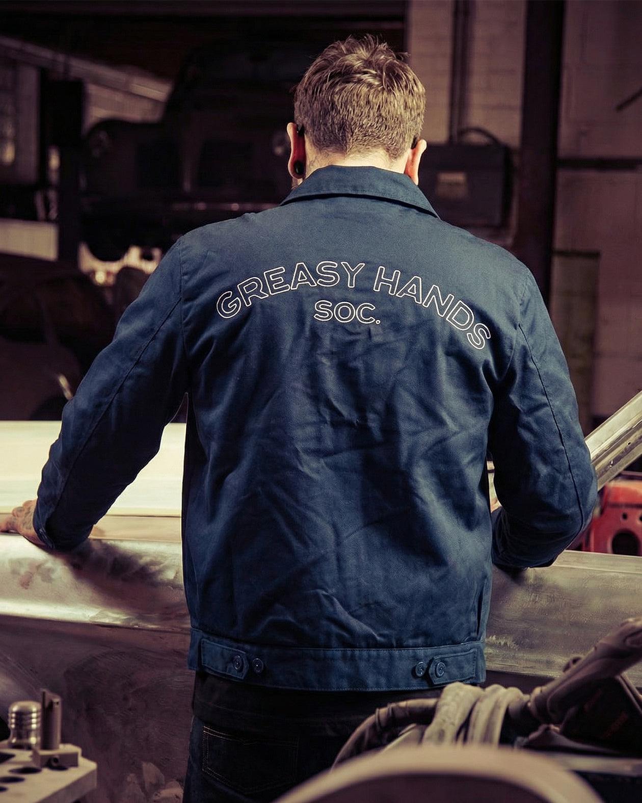 Person wearing a blue jacket with 'Greasy Hands Soc.' text in a workshop setting
