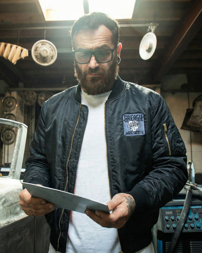 Man wearing a black flight jacket with a logo, holding a tablet in a workshop setting - Black Flight Jacket by Greasy Hands Society