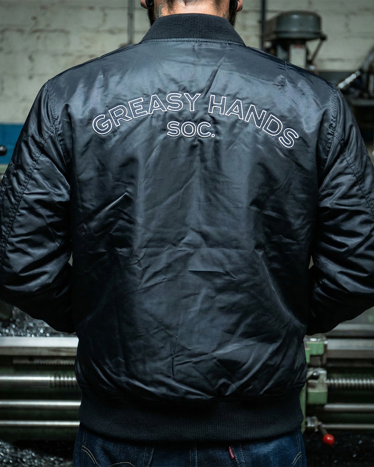 Person wearing a black flight jacket with 'Greasy Hands Soc.' text on the back, standing in an industrial setting.